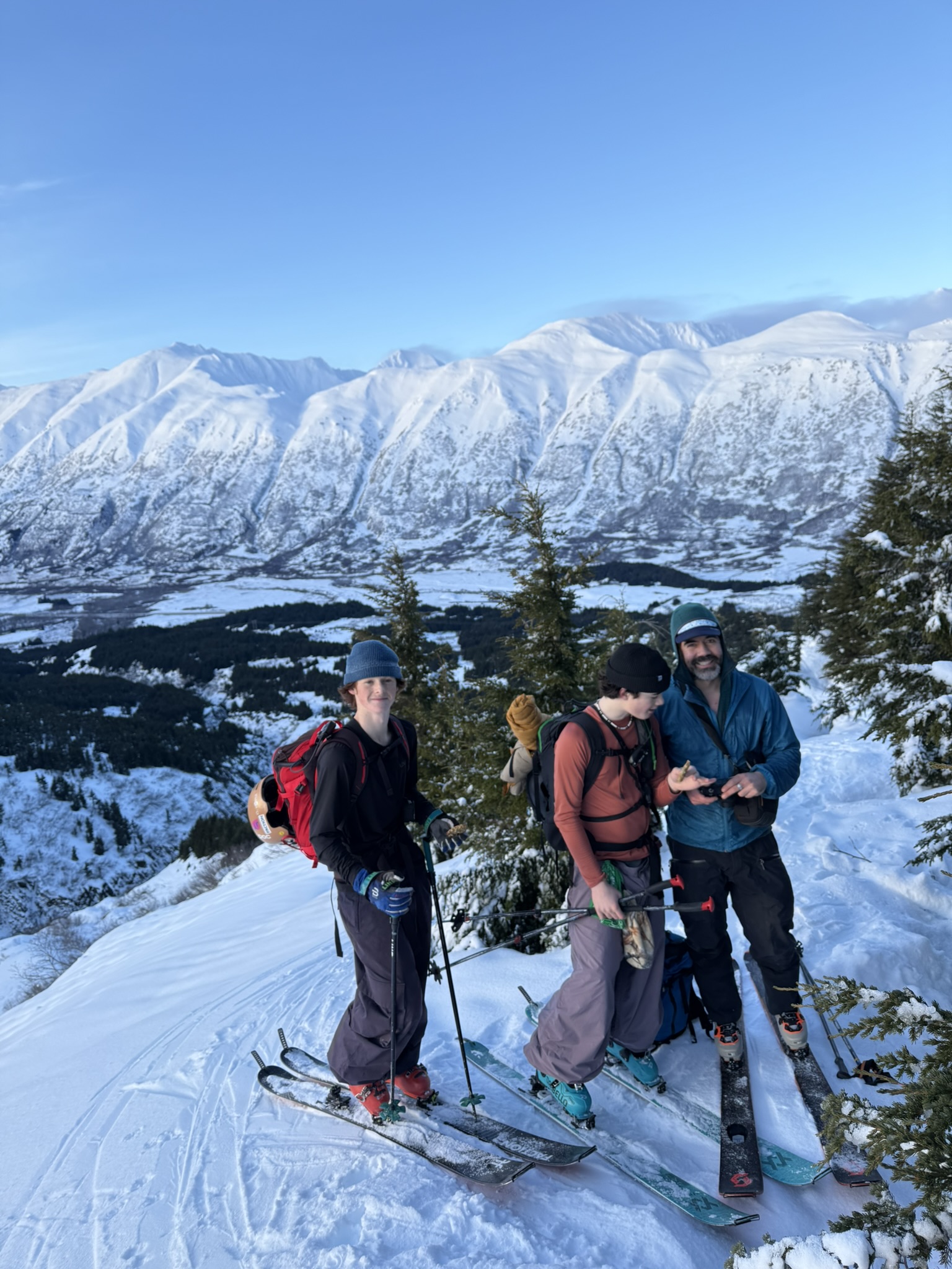 Three ski tourers on a snowy ridge above a valley, with Alaska's mountains behind them