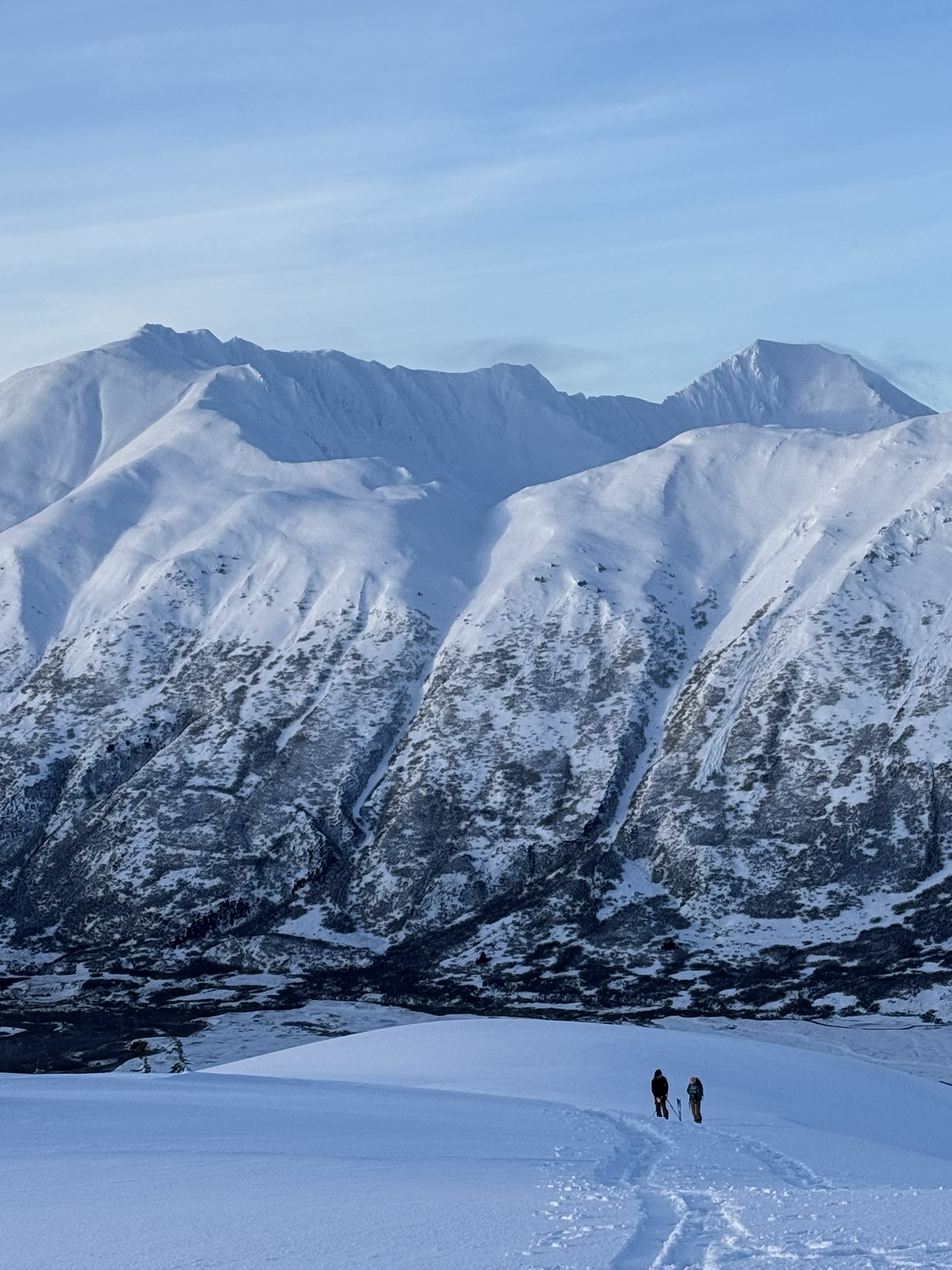 Two skiers skinning up a snowy slope toward big Alaska peaks