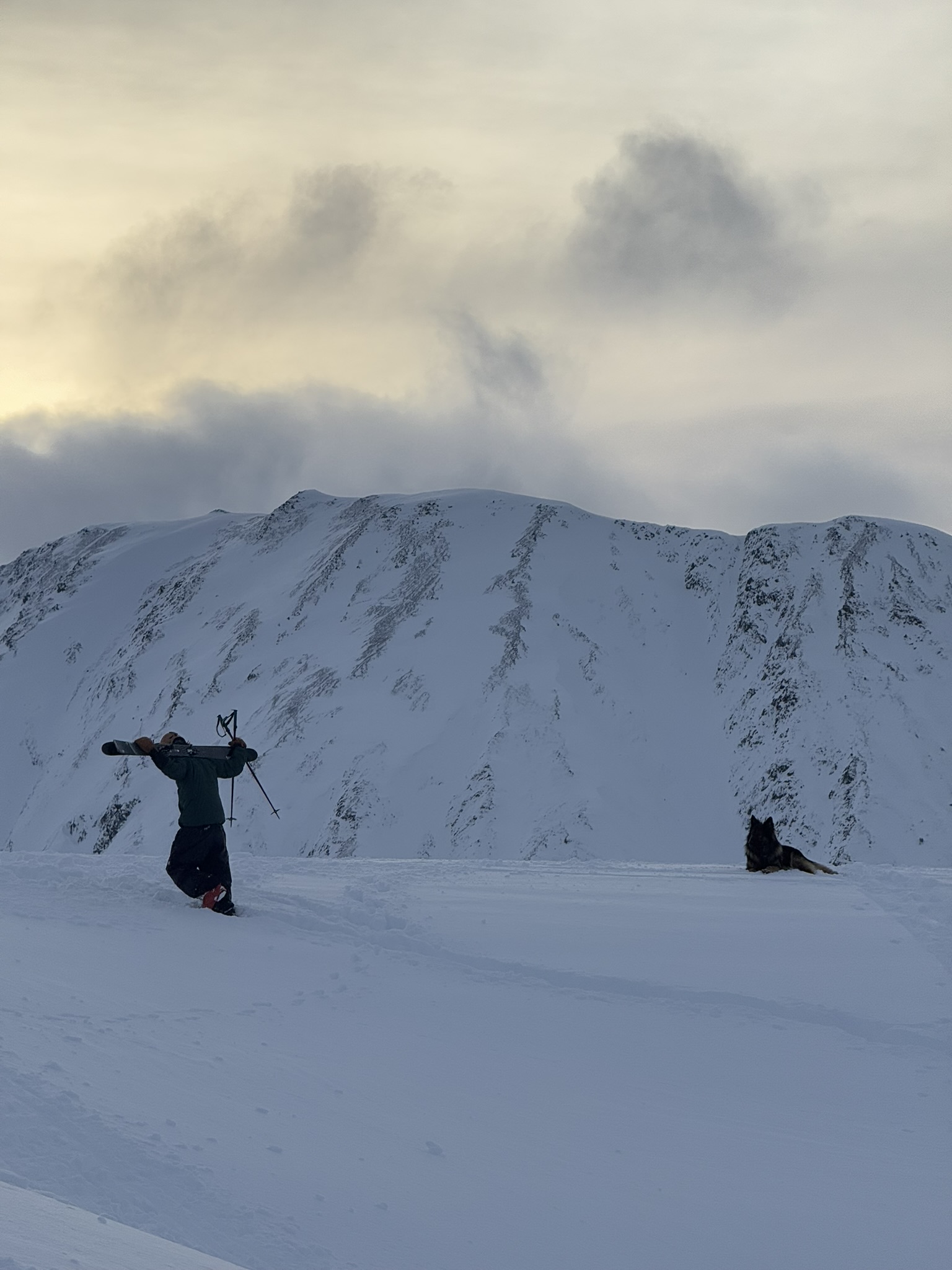 A skier carrying their skis along a snowy ridge under low cloud