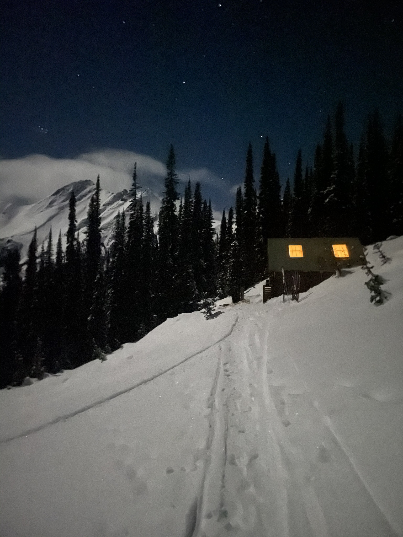 The skin track leading up to the cabin at night, its windows lit, under the stars