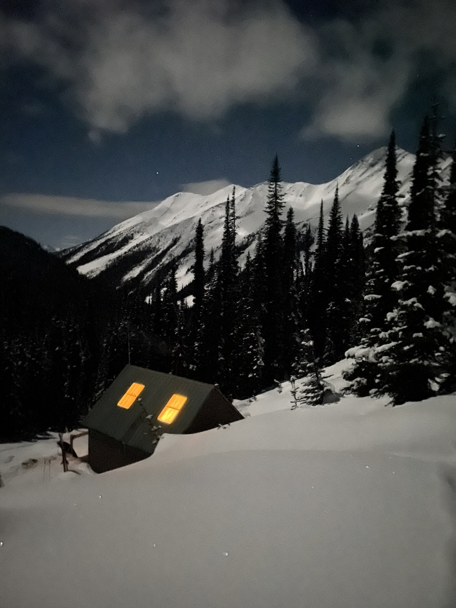 The cabin half buried in snow at night, its windows lit, with a moonlit peak behind it